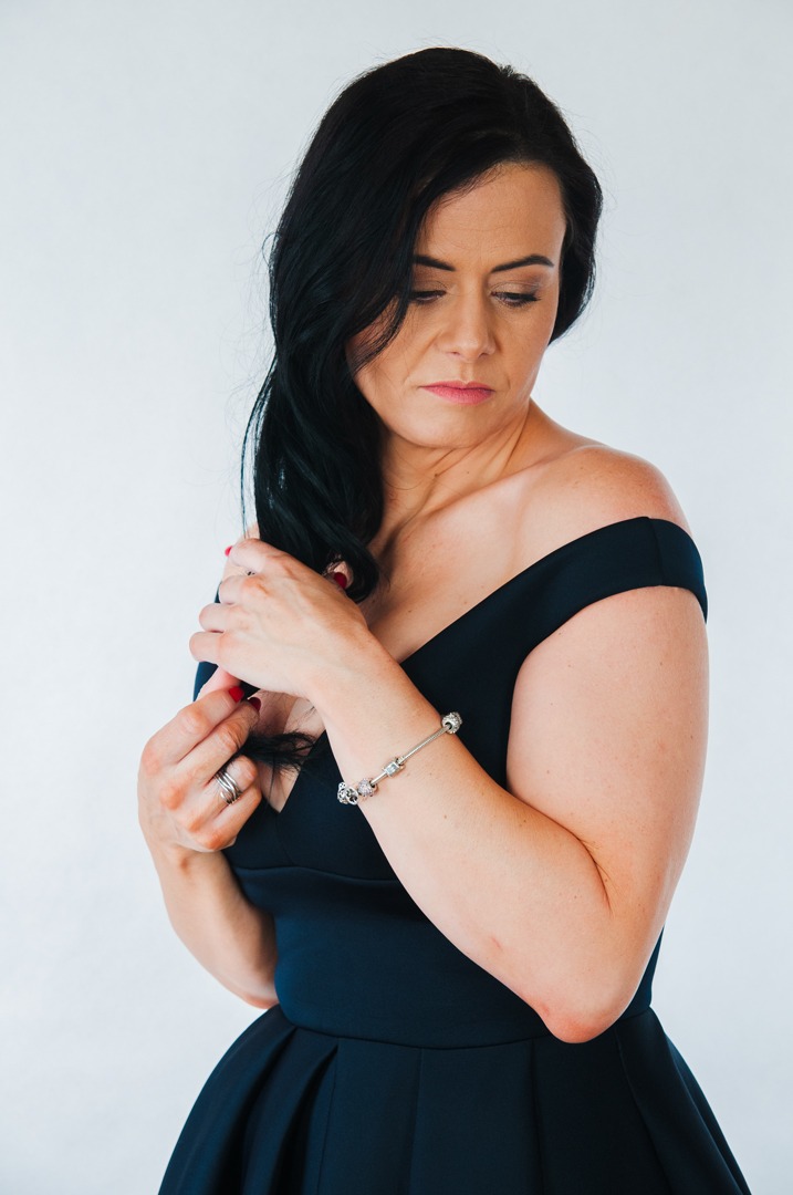 Kobieta w granatowej sukience z odkrytymi ramionami, dotyka włosów, na nadgarstku srebrna bransoletka z charmsami, spogląda w dół.