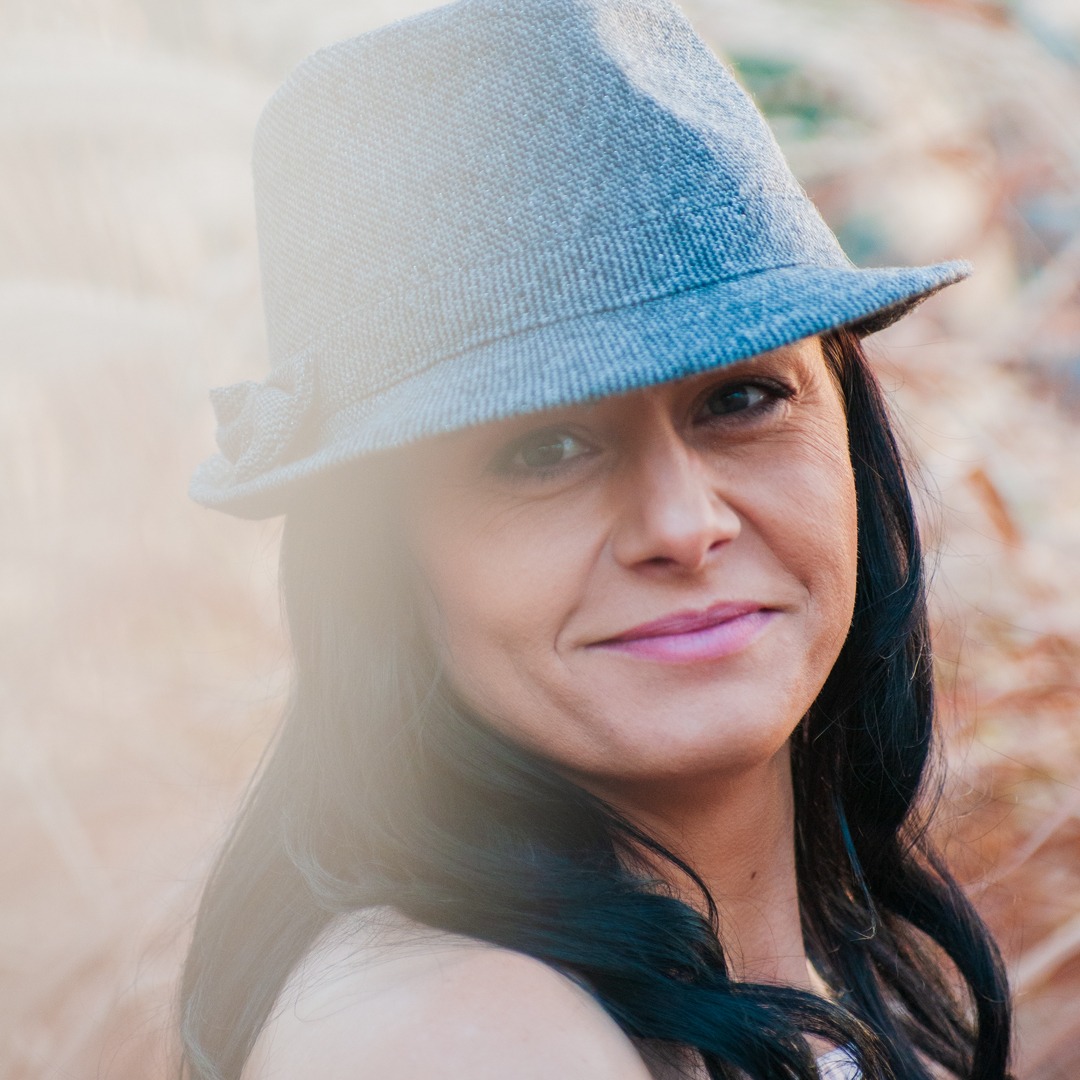 Portret kobiety w szarym kapeluszu z kokardą, czarne włosy, różowe usta, rozmyte tło w odcieniach brązu.