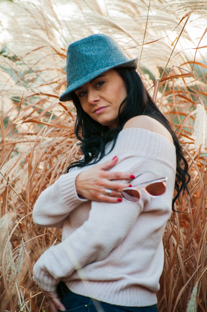 Kobieta w szarym kapeluszu i różowym swetrze pozuje na tle wysokich traw, trzymając okulary przeciwsłoneczne na ramieniu.
