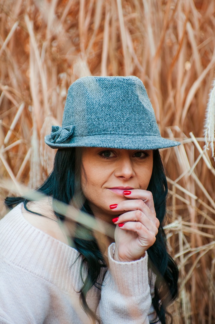 Portret kobiety w szarym kapeluszu z kokardą, z czerwonymi paznokciami, na tle suchej trawy.