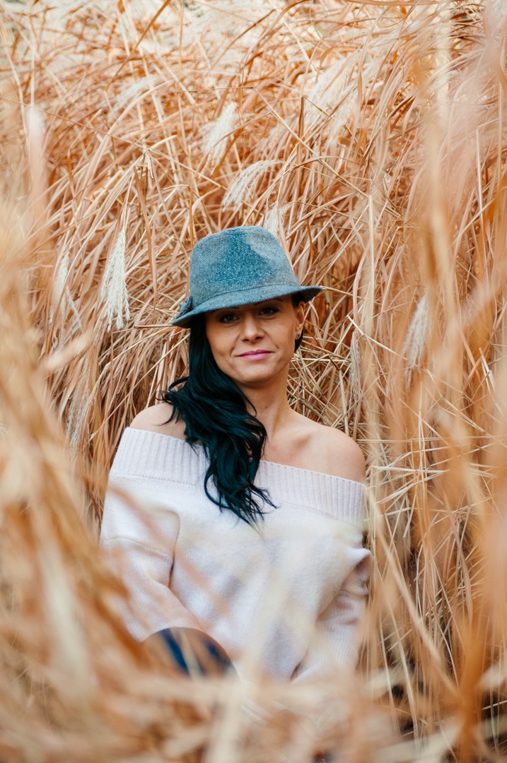 Portret kobiety w szarym kapeluszu i kremowym swetrze na tle wysokich traw w odcieniach beżu i brązu.
