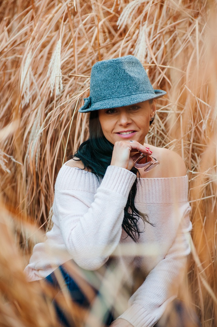 Kobieta w szarym kapeluszu i różowym swetrze pozuje na tle wysokich traw, trzymając okulary przeciwsłoneczne