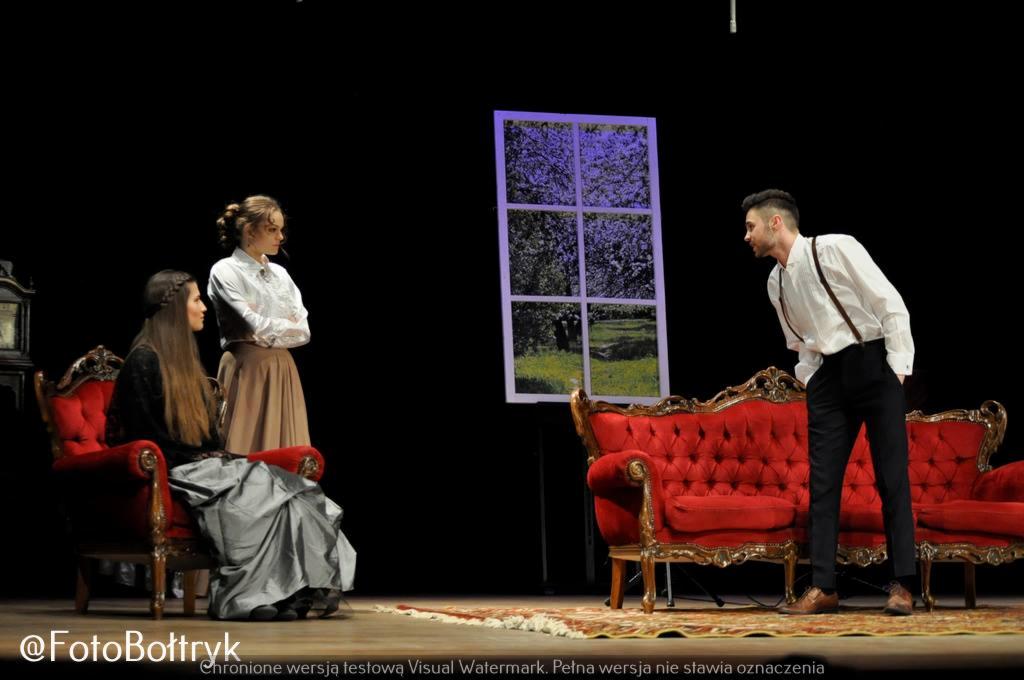 Scena teatralna: aktorzy w kostiumach na tle dekoracji okna i mebli w stylu retro.