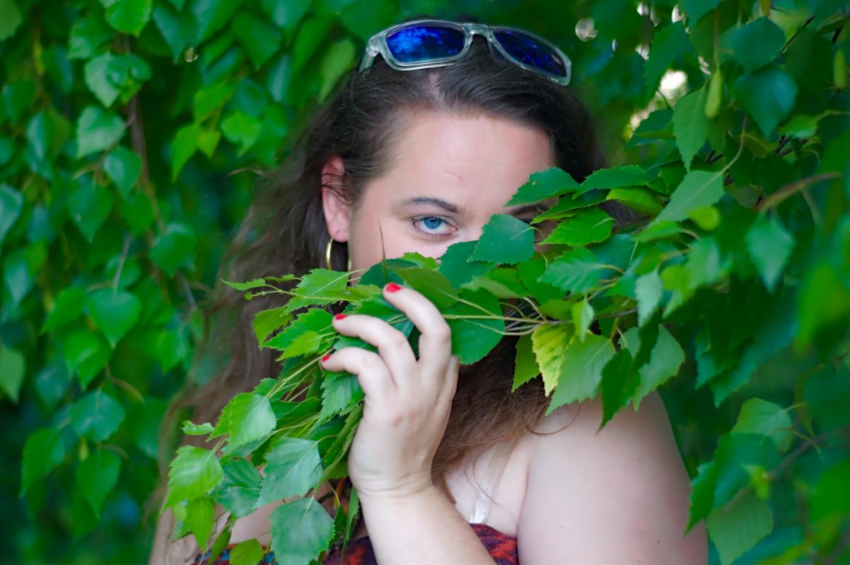 Portret kobiety z niebieskimi oczami, częściowo zasłoniętej gałązkami brzozy, zrobiony w Warszawie, z okularami przeciwsłonecznymi na głowie.