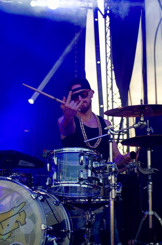 Perkusista w czarnej koszulce, okularach przeciwsłonecznych i z grubym złotym łańcuchem, grający na perkusji na scenie w niebieskim oświetleniu, pokazujący środkowy palec i trzymający pałeczkę.