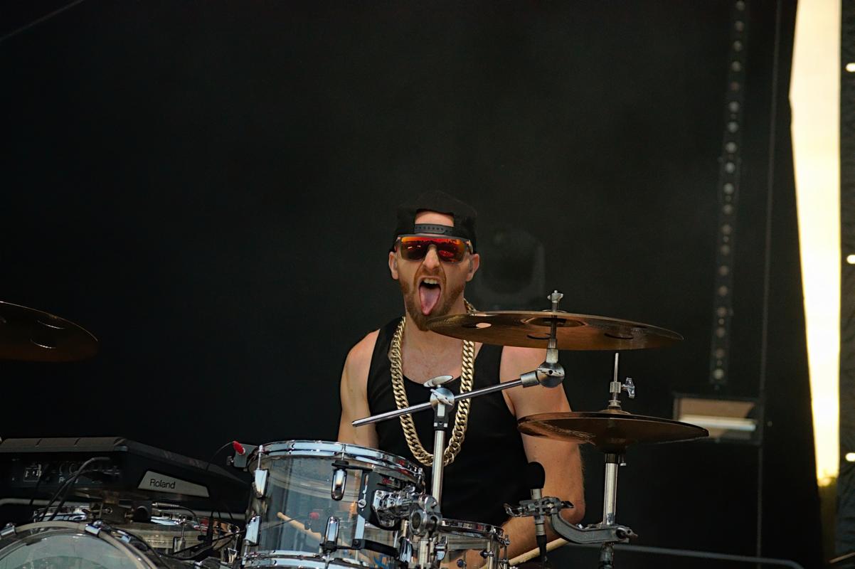 Perkusista w czapce i okularach, z wyciągniętym językiem, grający na perkusji podczas występu na scenie.