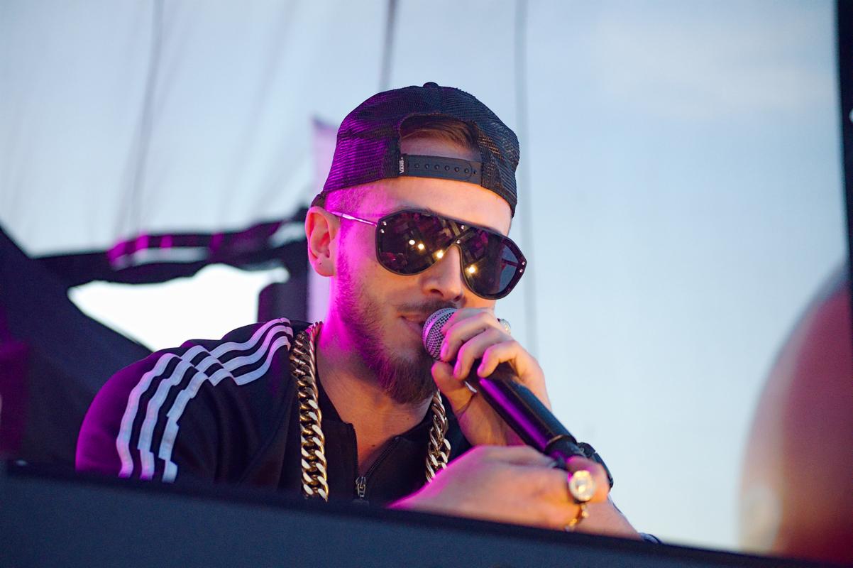 Mężczyzna z brodą w czapce z daszkiem i dużych okularach przeciwsłonecznych śpiewa do mikrofonu, ubrany w bluzę z trzema paskami i gruby złoty łańcuch.