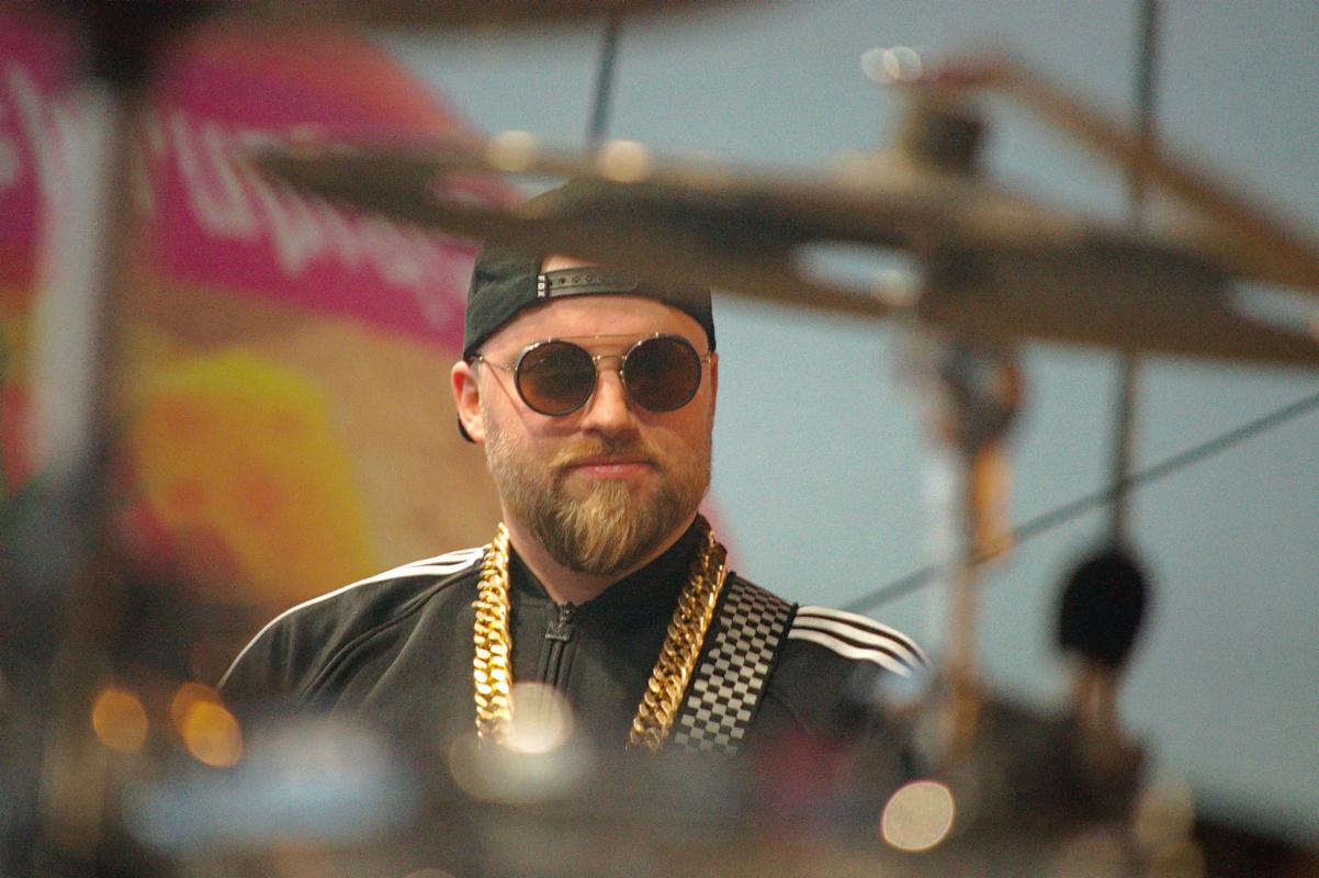 Mężczyzna z brodą i okularach przeciwsłonecznych, w czarnej bluzie z białymi paskami i złotym łańcuchem, częściowo zasłonięty elementami perkusji.