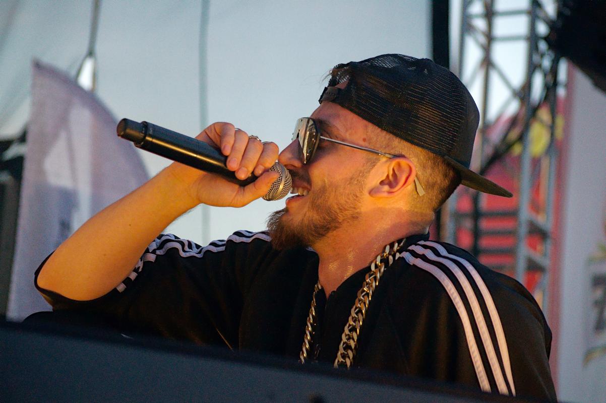Raper z brodą w czapce i okularach, śpiewa do mikrofonu na scenie podczas koncertu, widoczny łańcuch na szyi i pot na skórze.