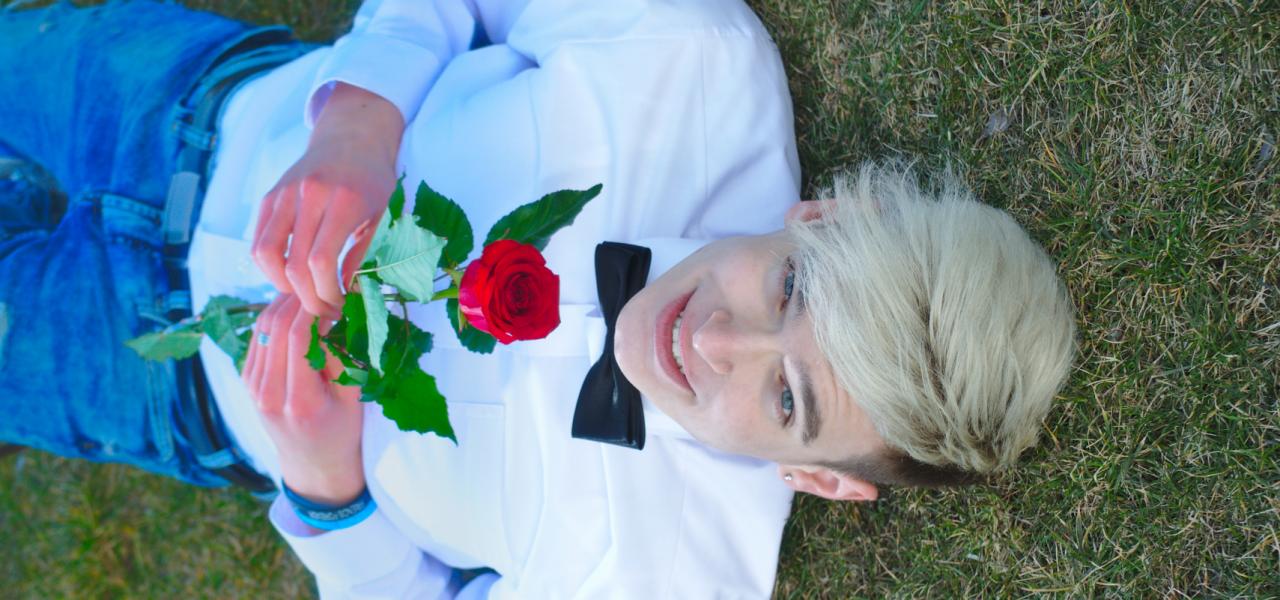 Młody mężczyzna z blond włosami i muszką, leżący na trawie, trzyma czerwoną różę, uśmiechając się do kamery.