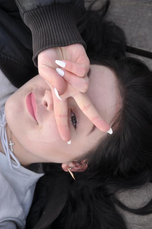 Portret młodej kobiety leżącej na ziemi, z ręką ułożoną w znak 'V' przed okiem, z widocznymi białymi paznokciami, delikatnym makijażem, srebrnym krzyżykiem na szyi i złotym kolczykiem w uchu.