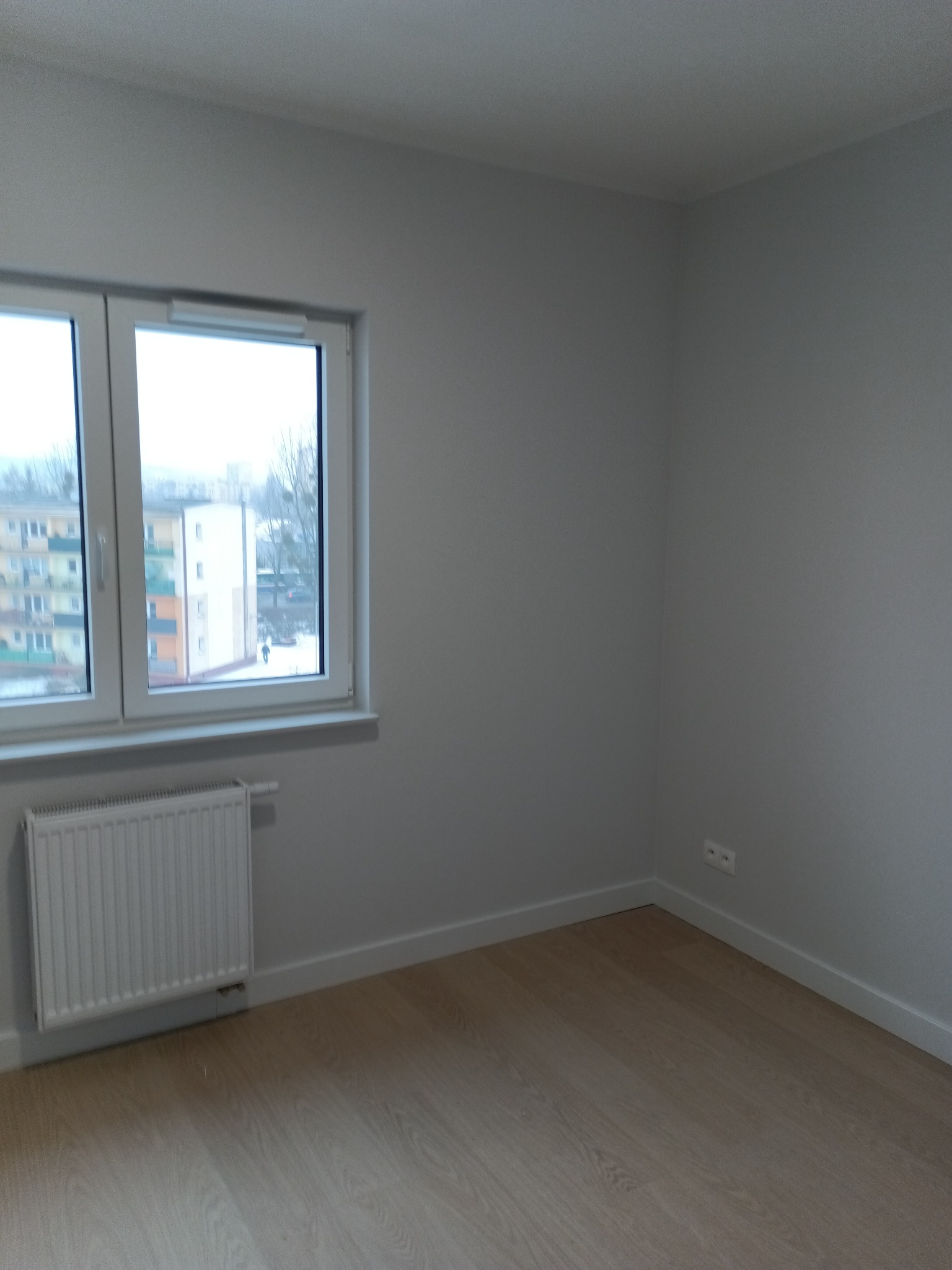 Jasne wnętrze pokoju po remoncie: świeżo pomalowane ściany w odcieniach szarości, panele podłogowe w kolorze drewna, białe listwy przypodłogowe i grzejnik pod oknem.