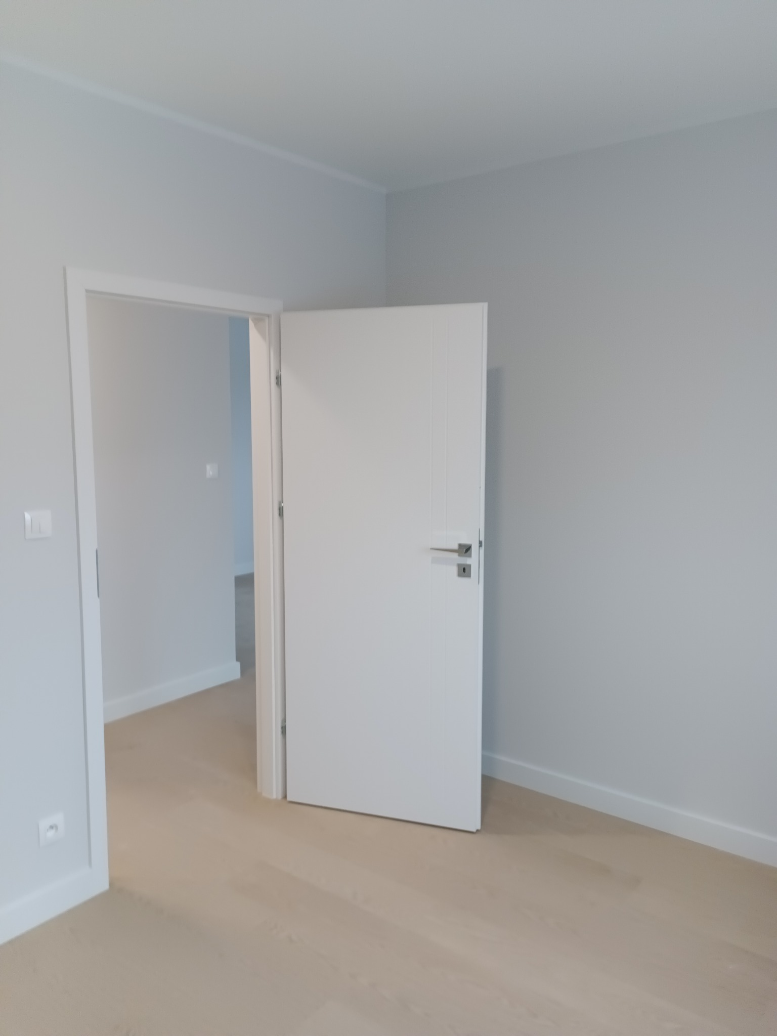 Jasne wnętrze po remoncie: białe ściany, drewniana podłoga i częściowo otwarte białe drzwi z srebrną klamką. Widoczny fragment listwy przypodłogowej i gniazdka elektrycznego.