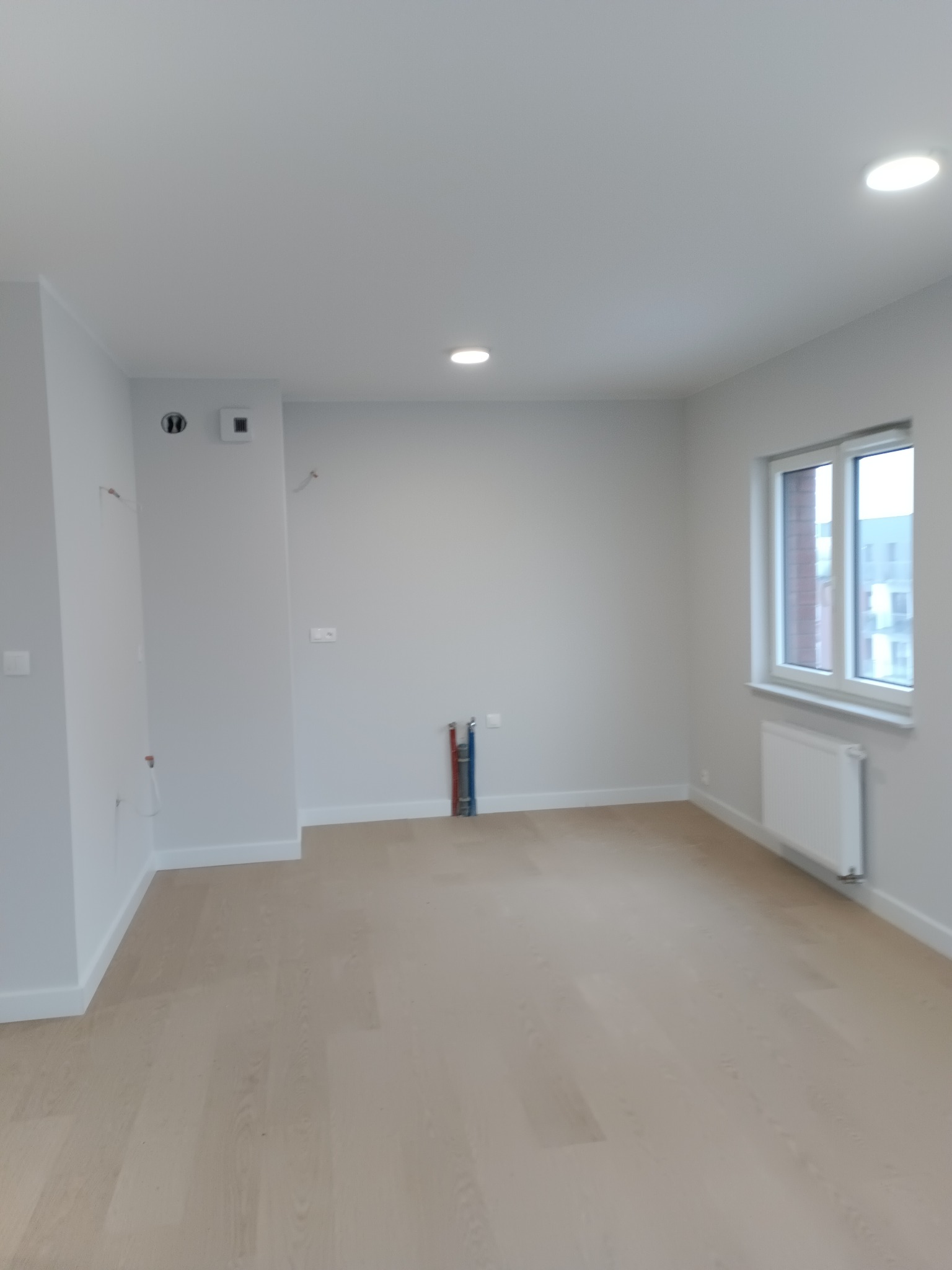 Jasne panele podłogowe w nowym, pomalowanym na szaro pokoju z grzejnikiem pod oknem. Widoczne piony instalacji. Minimalistyczne wykończenie.