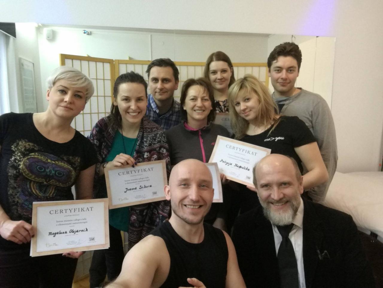 Grupa osób pozuje do zdjęcia, trzymając certyfikaty ukończenia kursu masażu i terapii naturalnych. Widoczny mężczyzna w garniturze i kilka osób w strojach codziennych, w tle parawan.