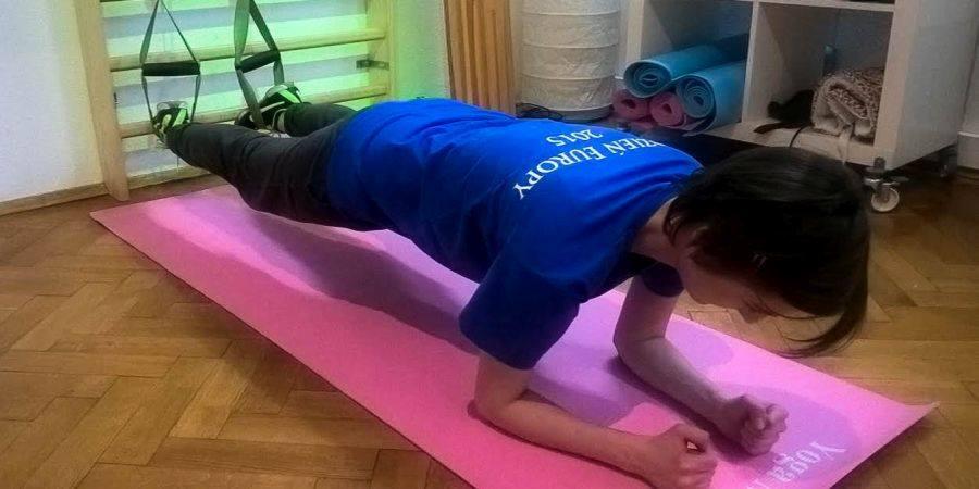 Kobieta w niebieskiej koszulce wykonuje ćwiczenie plank z nogami zawieszonymi na taśmach TRX, leżąc na różowej macie do jogi w pomieszczeniu z drewnianą podłogą.