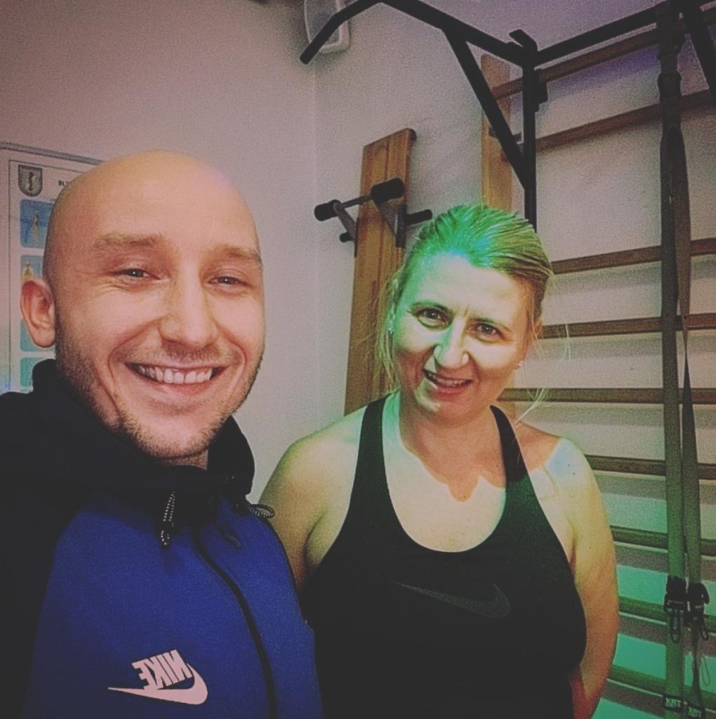 Uśmiechnięty mężczyzna w niebieskiej bluzie Nike i kobieta w czarnym topie na tle ściany z drabinką gimnastyczną i plakatem.