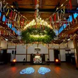 Dekoracja sali weselnej z oświetleniem: girlandy, wstążki, reflektory i DJ-ka. W centralnym punkcie żyrandol opleciony zielenią. Parkiet z projekcjami świetlnymi.