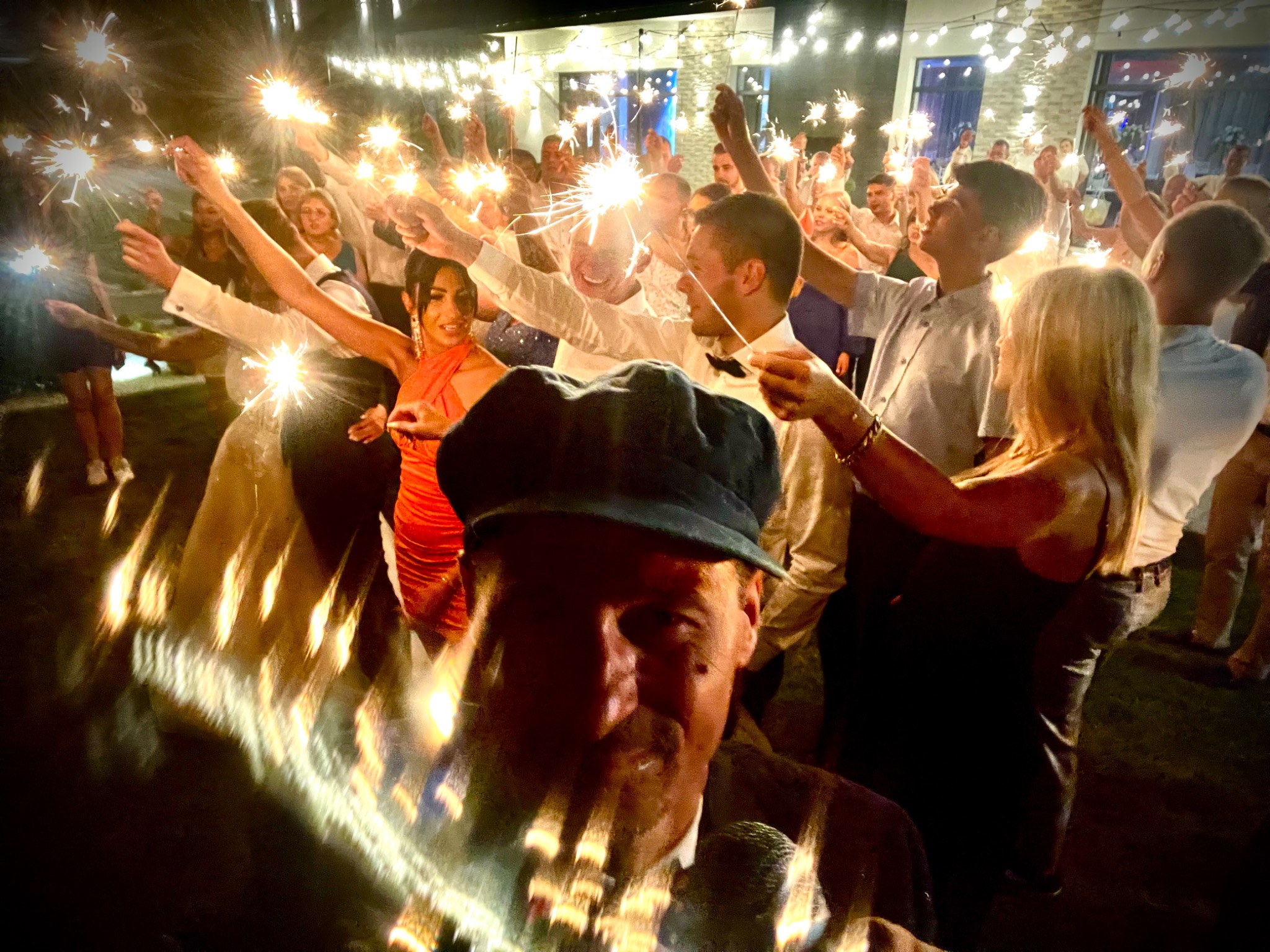 Rozświetlona uroczystość: grupa osób trzymająca zimne ognie przed budynkiem z dekoracyjnym oświetleniem. W pierwszym planie mężczyzna z mikrofonem.