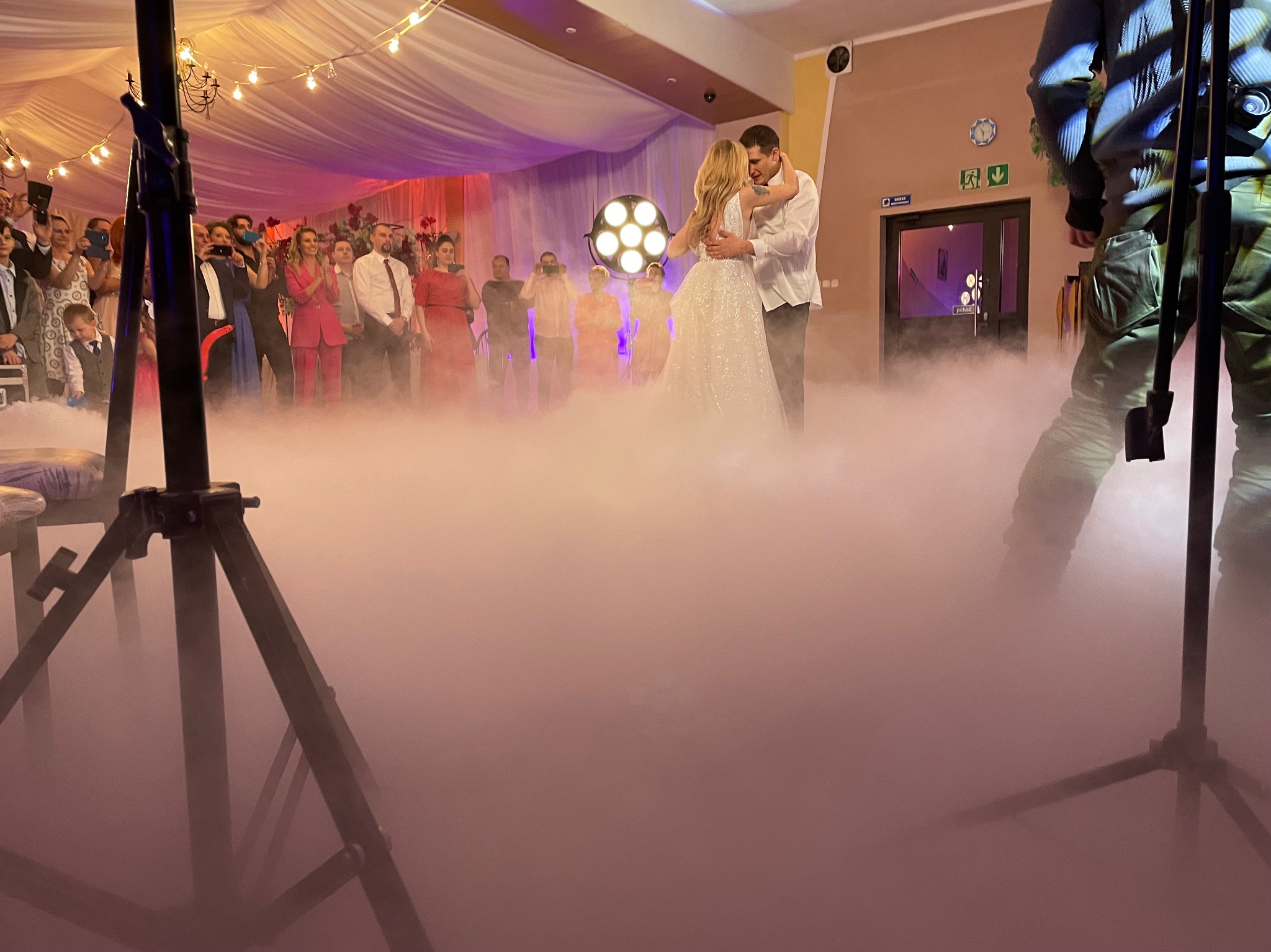 Para młoda w tańcu w chmurze dymu na tle gości i oświetlenia scenicznego. Widoczny statyw oświetleniowy. Dekoracja sali w stylu glamour.