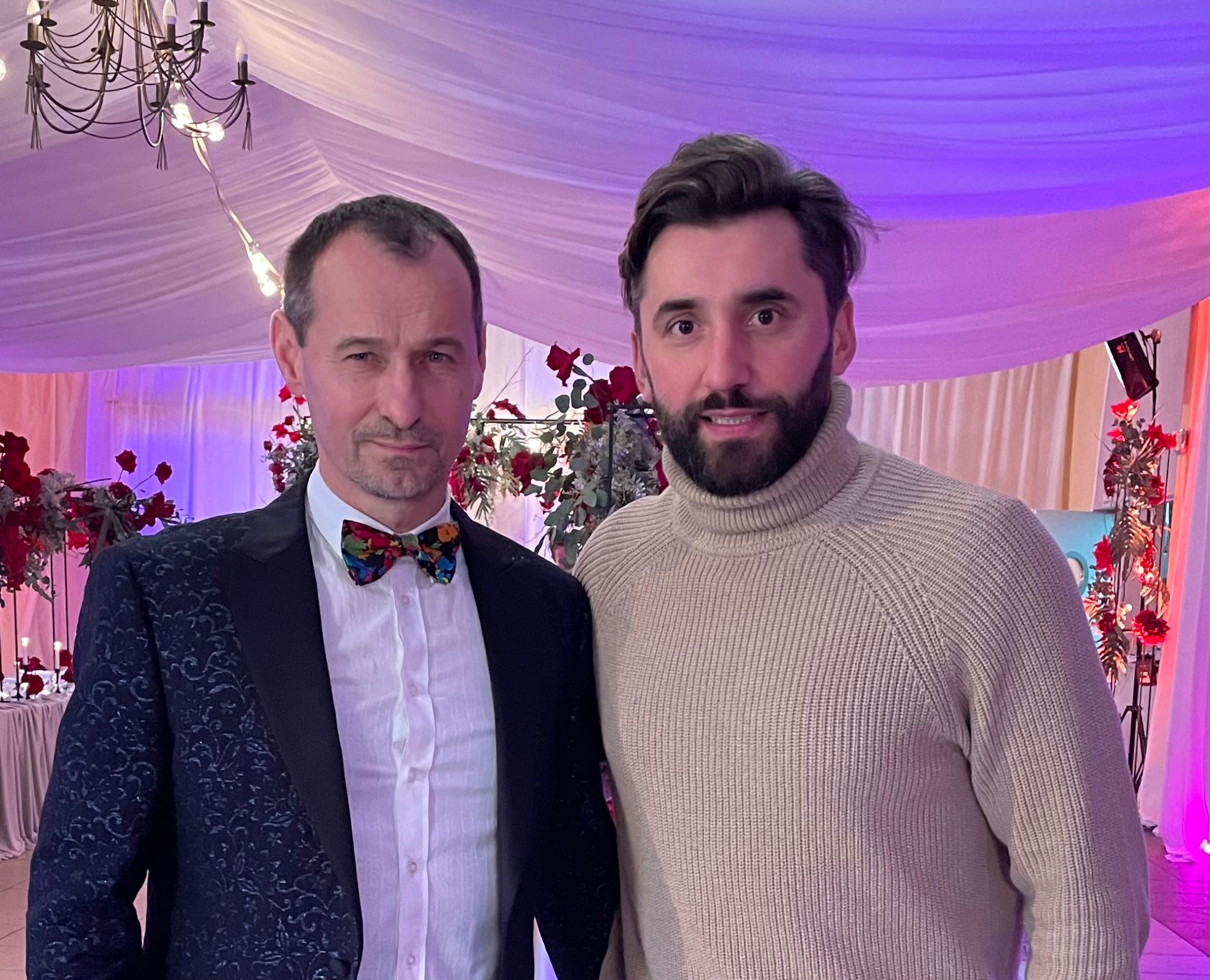 Portret dwóch mężczyzn na tle dekoracji kwiatowych i oświetlenia scenicznego, prawdopodobnie podczas wydarzenia muzycznego lub uroczystości.