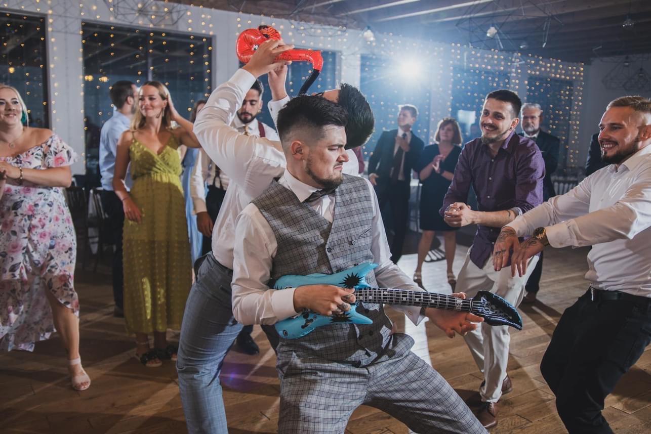 Rozbawiony zespół muzyczny z dmuchanymi gitarami i bokserkami podczas występu na parkiecie, oświetlonym dekoracyjnymi lampkami.