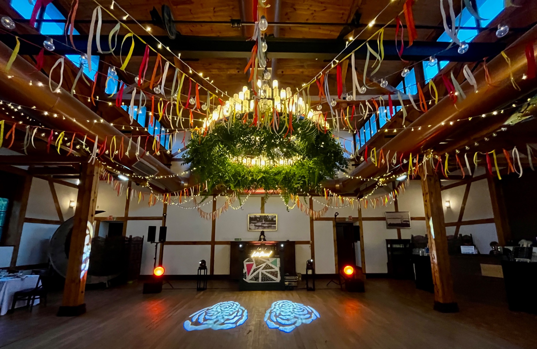 Dekoracja sali weselnej z oświetleniem: girlandy, wstążki, reflektory i DJ-ka. W centralnym punkcie żyrandol opleciony zielenią. Parkiet z projekcjami świetlnymi.