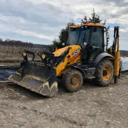 Żółta koparko-ładowarka JCB na nierównym, kamienistym terenie, z widocznym łyżką i ramieniem kopiącym, w tle drzewa i pochmurne niebo.