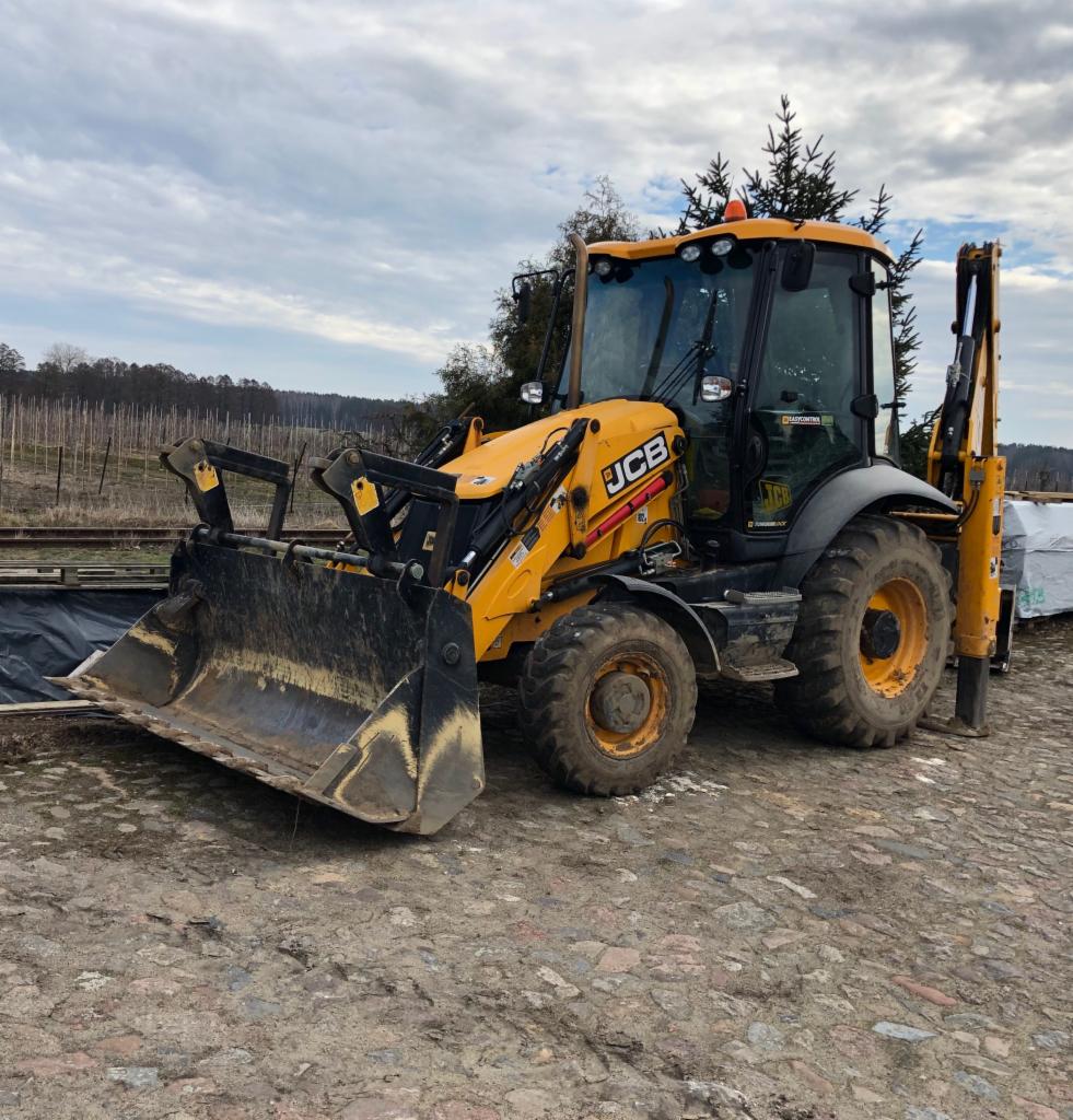 Żółta koparko-ładowarka JCB na nierównym, kamienistym terenie, z widocznym łyżką i ramieniem kopiącym, w tle drzewa i pochmurne niebo.