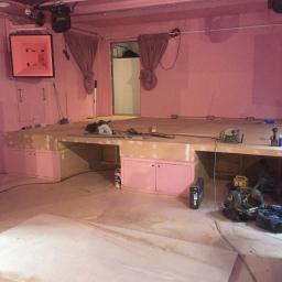 Dominik Spychała - Wnętrze lokalu z podwyższoną drewnianą sceną w trakcie budowy, różowe ściany, widoczne narzędzia i materiały budowlane, surowe wykończenie.