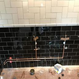 Dominik Spychała - Instalacja miedziana w łazience podczas remontu, widoczne czarne i białe płytki na ścianach, narzędzia i elementy instalacyjne na podłodze.