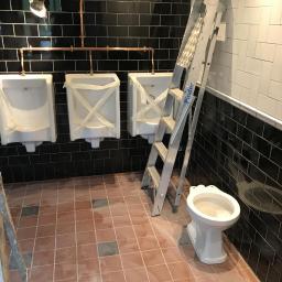 Dominik Spychała - Publiczna toaleta w trakcie prac wykończeniowych: trzy pisuary zabezpieczone folią, częściowo zamontowana toaleta, drabina oparta o ścianę z czarnych płytek.