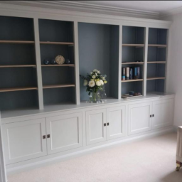 Dominik Spychała - Jasnoszara zabudowa meblowa z szafkami i półkami, dekorowana bukietem kwiatów w wazonie i zegarem, na tle ściany w odcieniach szarości.