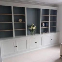 Jasnoszara zabudowa meblowa z szafkami i półkami, dekorowana bukietem kwiatów w wazonie i zegarem, na tle ściany w odcieniach szarości.