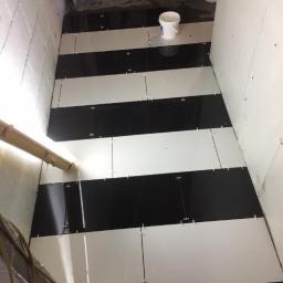 Dominik Spychała - Układanie czarno-białych płytek podłogowych w pomieszczeniu; widoczne krzyżyki dystansowe i wiadro z zaprawą.