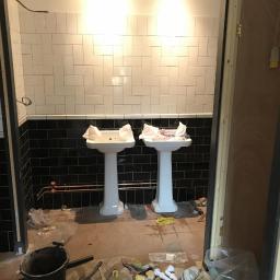 Dominik Spychała - Łazienka w trakcie prac remontowych: dwie białe umywalki na postumentach, częściowo wyłożone czarno-białe płytki, widoczne narzędzia i materiały budowlane na podłodze.