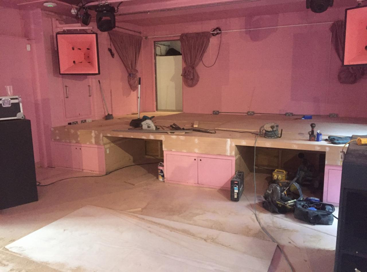 Wnętrze lokalu z podwyższoną drewnianą sceną w trakcie budowy, różowe ściany, widoczne narzędzia i materiały budowlane, surowe wykończenie.