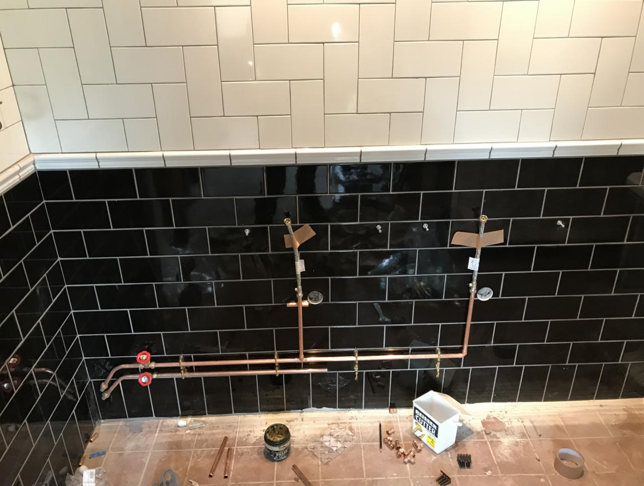 Instalacja miedziana w łazience podczas remontu, widoczne czarne i białe płytki na ścianach, narzędzia i elementy instalacyjne na podłodze.