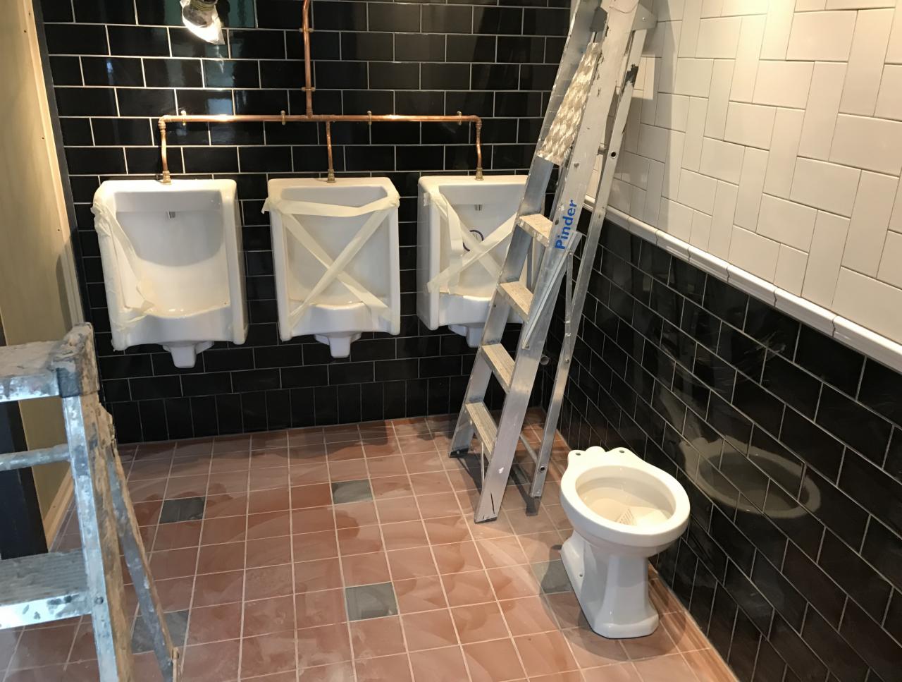 Publiczna toaleta w trakcie prac wykończeniowych: trzy pisuary zabezpieczone folią, częściowo zamontowana toaleta, drabina oparta o ścianę z czarnych płytek.