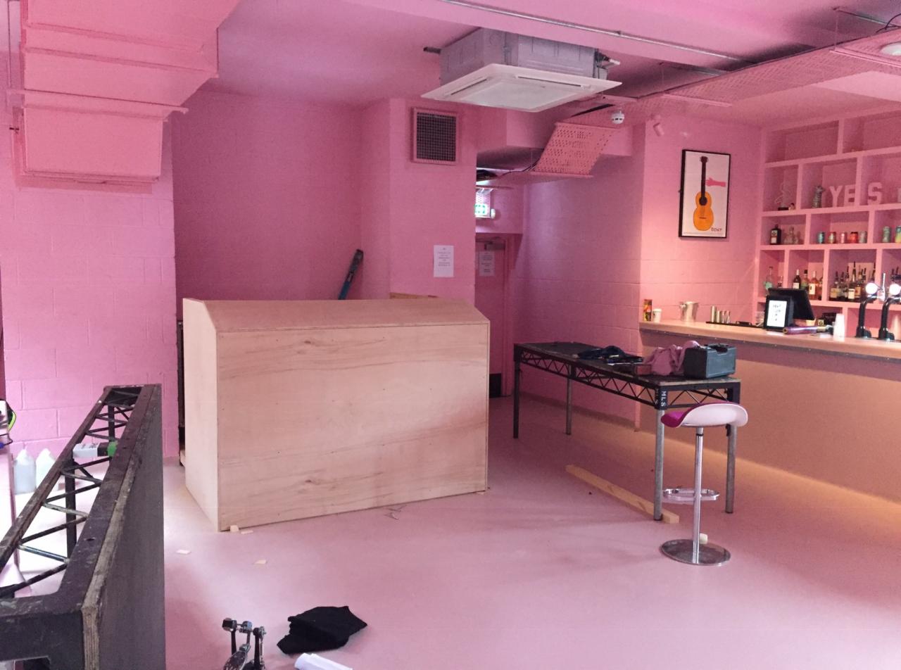 Wnętrze lokalu w całości pomalowane na różowo, z widocznym barem, stolikiem, stołkiem barowym oraz niezabudowaną konstrukcją z płyty, prawdopodobnie w trakcie prac wykończeniowych.
