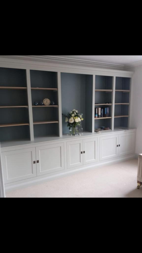 Jasnoszara zabudowa meblowa z szafkami i półkami, dekorowana bukietem kwiatów w wazonie i zegarem, na tle ściany w odcieniach szarości.