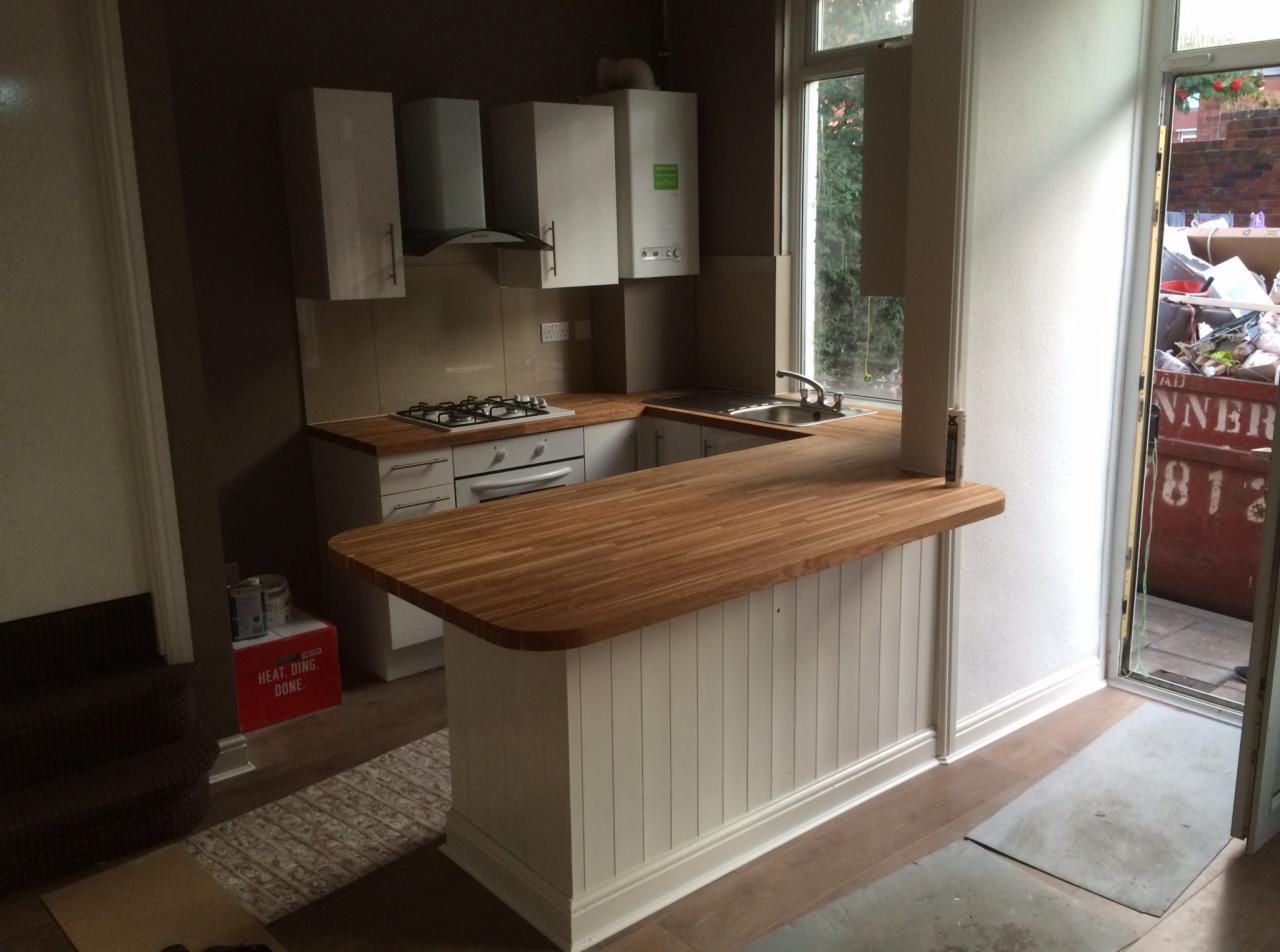 Kuchnia po remoncie z drewnianym blatem barowym, widok na zewnątrz na kontener na odpady budowlane, częściowo ułożona podłoga.