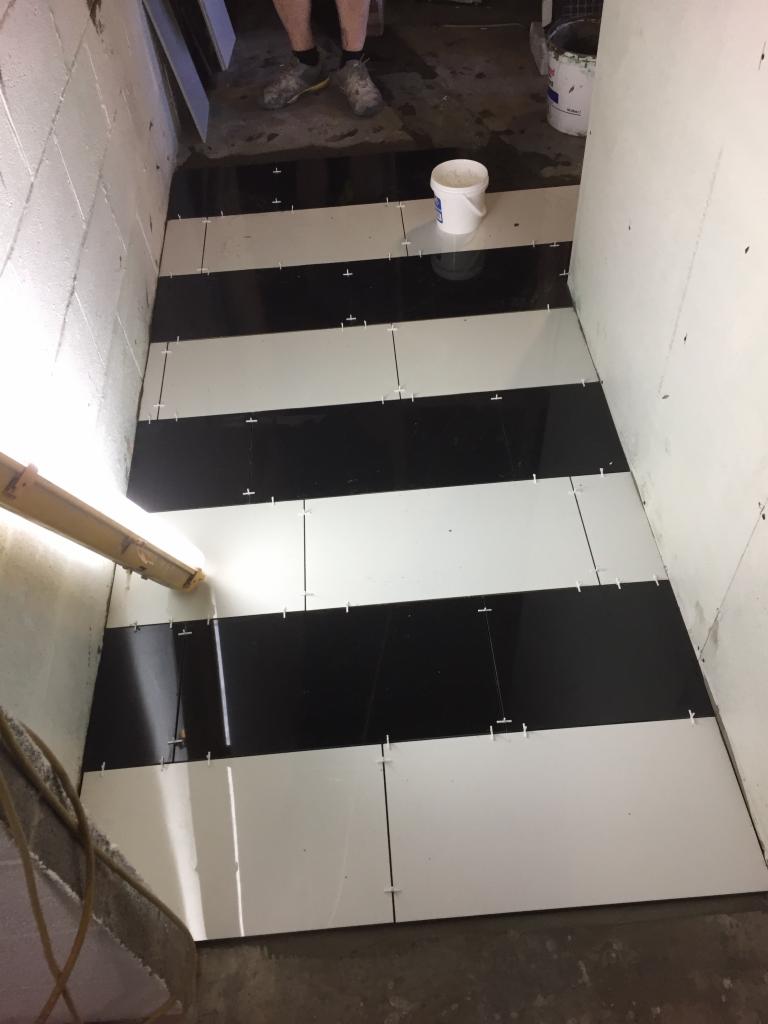 Układanie czarno-białych płytek podłogowych w pomieszczeniu; widoczne krzyżyki dystansowe i wiadro z zaprawą.