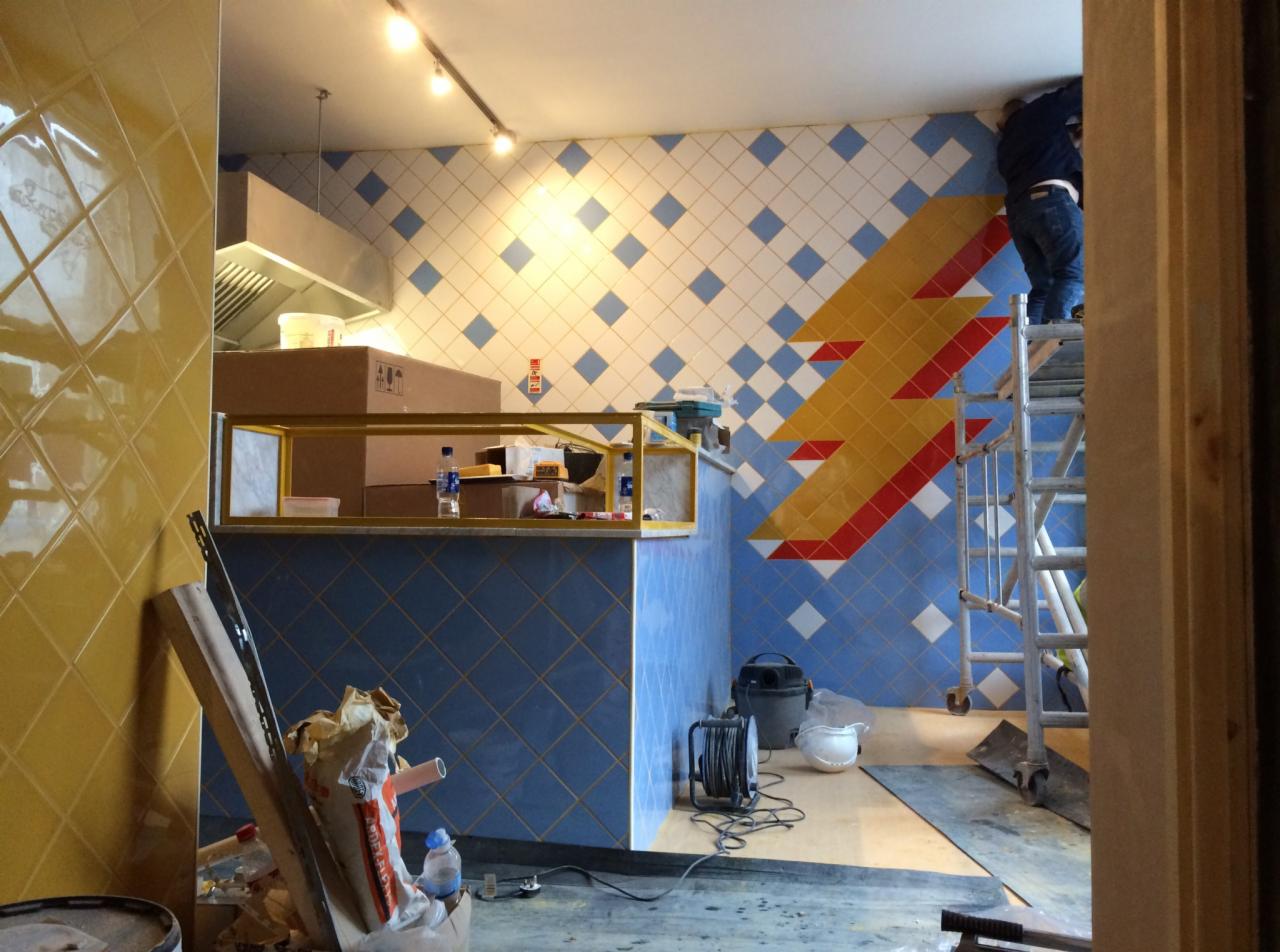 Wnętrze lokalu w trakcie wykańczania: układanie płytek z motywem błyskawicy na ścianie, widoczny pracownik na drabinie, niezabezpieczone podłoże, materiały budowlane i narzędzia.