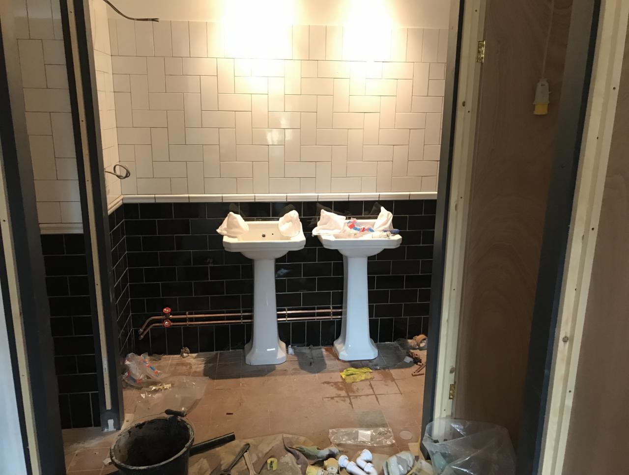 Łazienka w trakcie prac remontowych: dwie białe umywalki na postumentach, częściowo wyłożone czarno-białe płytki, widoczne narzędzia i materiały budowlane na podłodze.