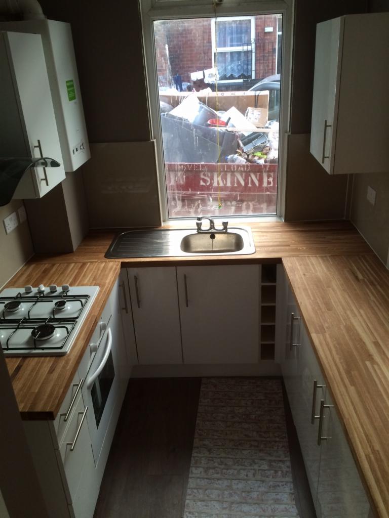 Wąska kuchnia z białymi szafkami, drewnianym blatem i widokiem na śmietnik za oknem.