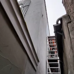 Dwóch pracowników na aluminiowej drabinie maluje wąską ścianę budynku między dwiema kamienicami, widok z dołu na pochmurne niebo.