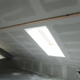 Poddasze ze skośnym sufitem i oknem dachowym w trakcie prac wykończeniowych, widoczne łączenia płyt i ślady szpachlowania.