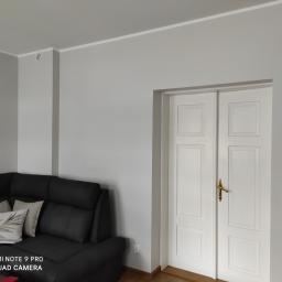 Firma Usługowa Martyna Kowal - Szare wnętrze po odświeżeniu: ściany pomalowane na jednolity szary kolor, białe dwuskrzydłowe drzwi z złotą klamką, fragment ciemnej sofy z poduszkami w rogu pokoju.