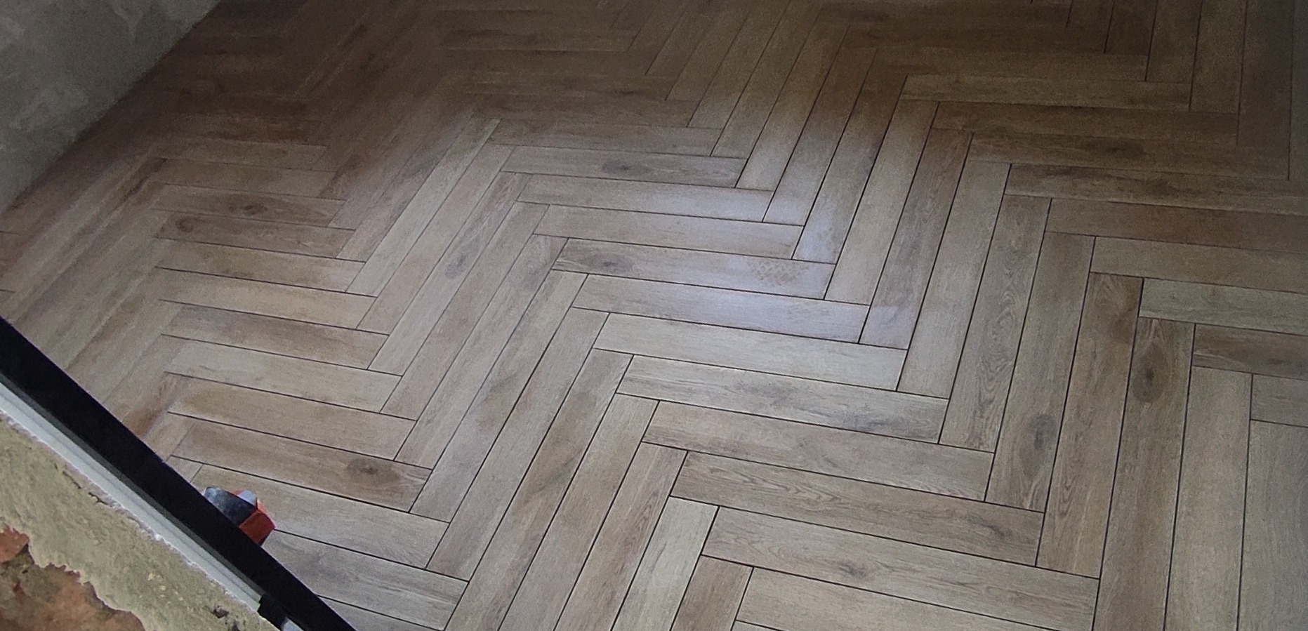 Ułożona w jodełkę podłoga z płytek imitujących drewno, widoczne niedokończone wykończenie ściany i listwy przypodłogowej.